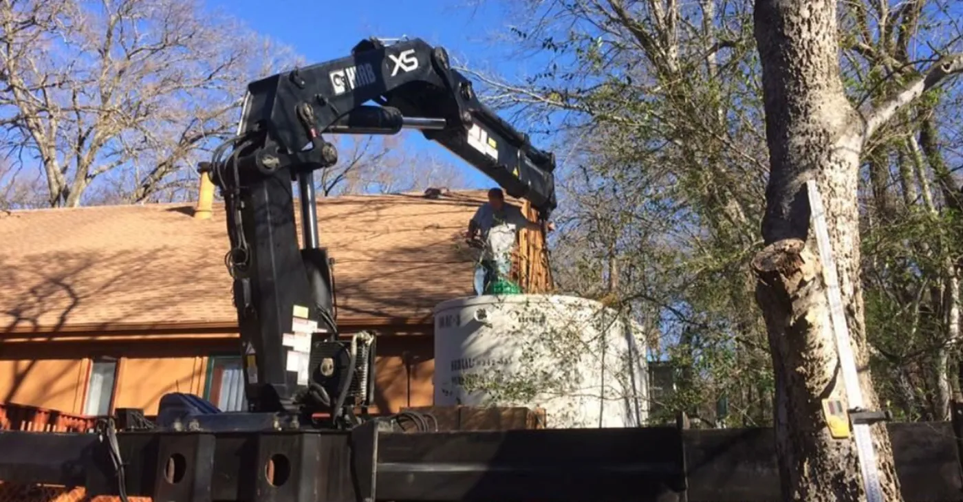 A boom truck lowering a new septic tank into place beside a brick house