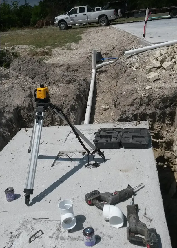 A laser level on a tripod set on a concrete tank lid, with PVC fittings and tools ready for an installation