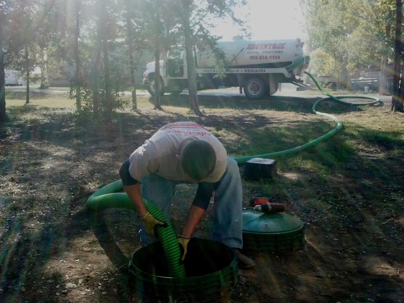 An Advantage Septic technician guiding a green vacuum hose into an open aerobic tank, the lettered pump truck parked behind him